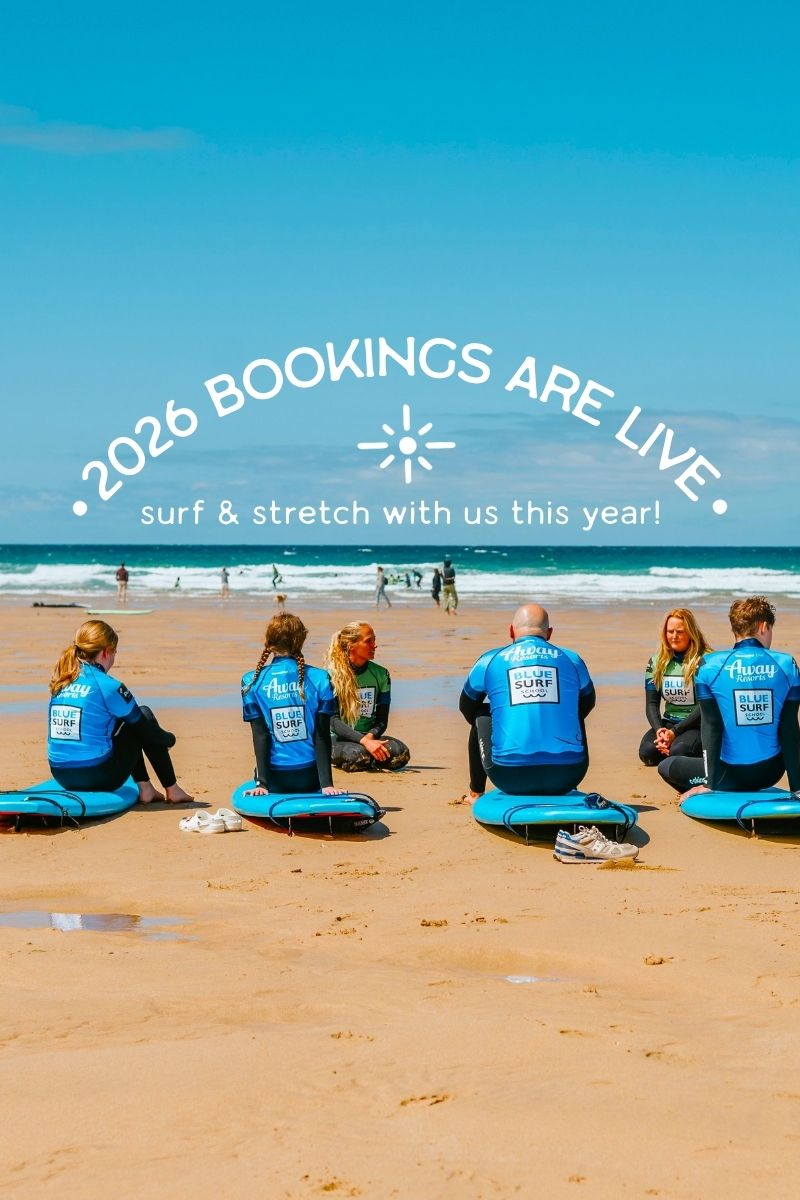 Group of people in Blue Surf tops sitting on surfboards on a surf lesson in Watergate Bay, Newquay, overlooking beautiful white sand and blue seas with text overlay about 2026 bookings being open.