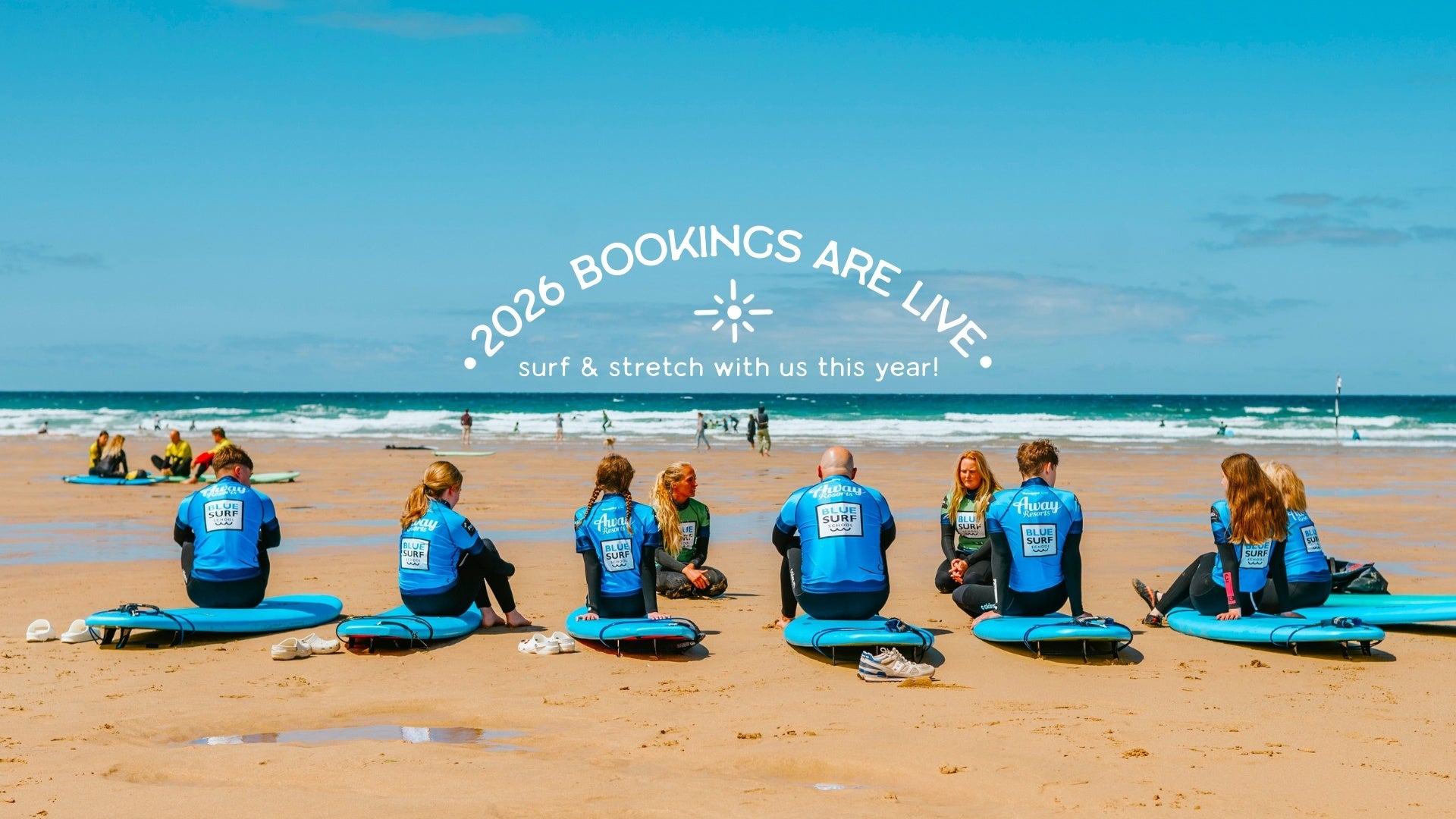 Group of people in Blue Surf tops sitting on surfboards on a surf lesson in Watergate Bay, Newquay, overlooking beautiful white sand and blue seas with text overlay about 2026 bookings being open.